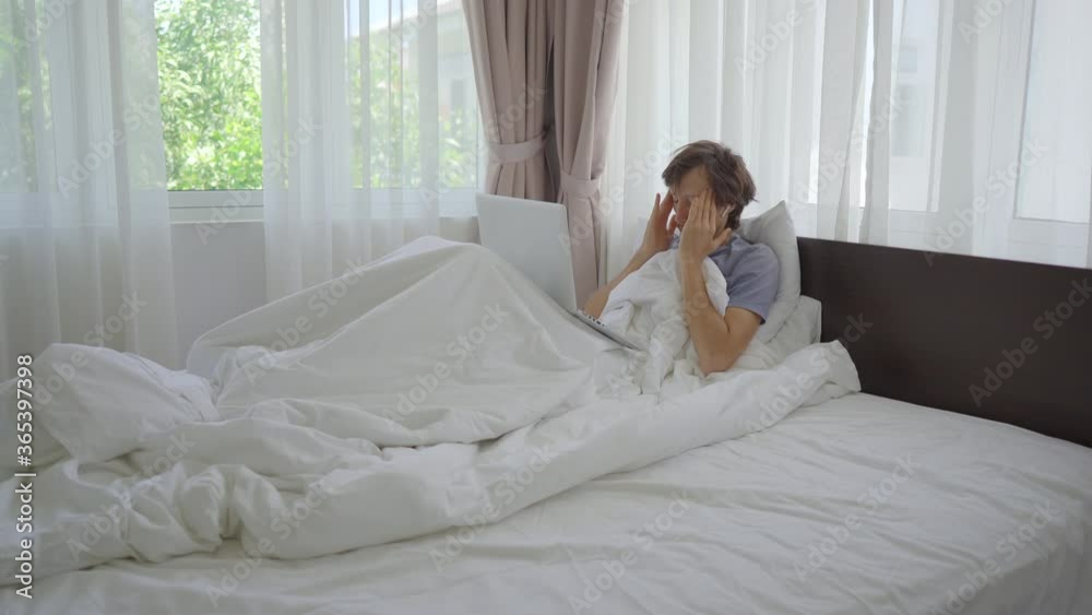 A sad young man in a bed talks with someone through video conferencing using a notebook. He is tired of sitting at home during social isolation A sad young man in a bed talks with someone through video conferencing using a notebook. He is tired of sitting at home during social isolation