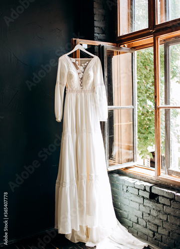 Modern wedding dress hanging at window in soft morning light. Stylish luxury wedding dress with lace floral pattern in light.