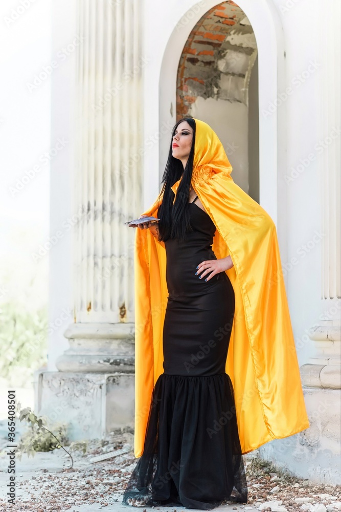 Naklejka premium Mystical woman in yellow cloak, black dress, and tarot cards at old town, Halloween date, party outfit 