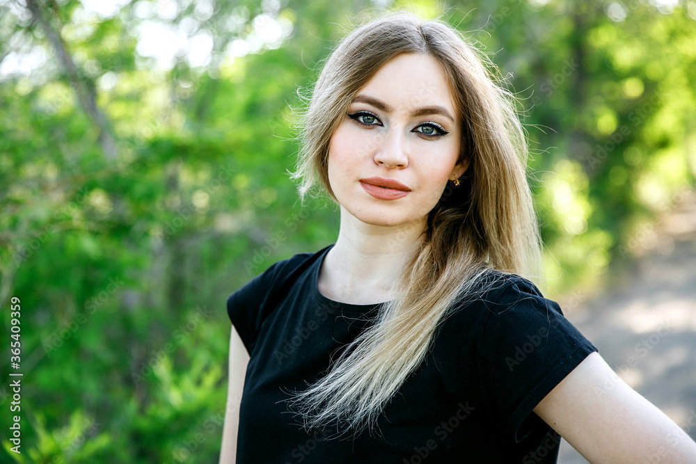 Outdoor portrait of a young pretty girl with long blond hair wearing a black t-shirt and blue jeans standing on the street against the background of green trees.