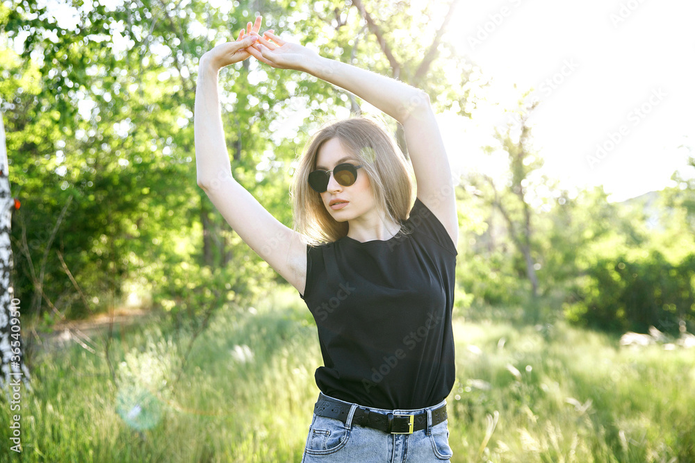 beautiful girl in sunglasses and sunglasses in a black T-shirt, arms raised up, looking towards the glare of the sun create a haze. summer