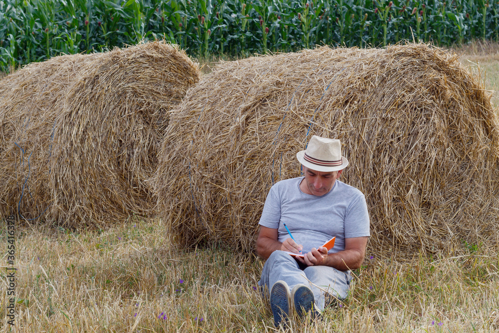 Farmer Writing in Notebook While leaning on Bale of Straw