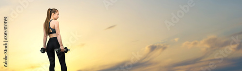 A young slim athletic girl in sportswear performs a set of exercises against the sunset by the sea. Performs exercises with dumbbells. Fitness and healthy lifestyle.