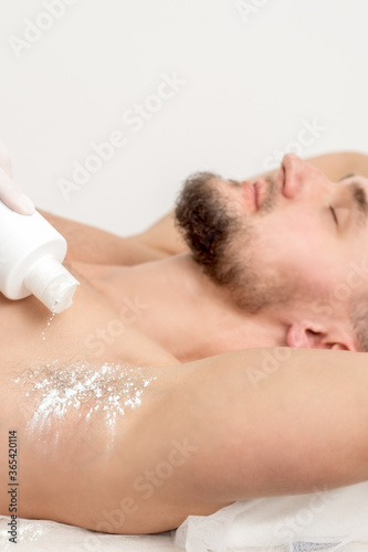 Hand of master depilation pouring talcum powder on armpit of young man before depilation procedure.
