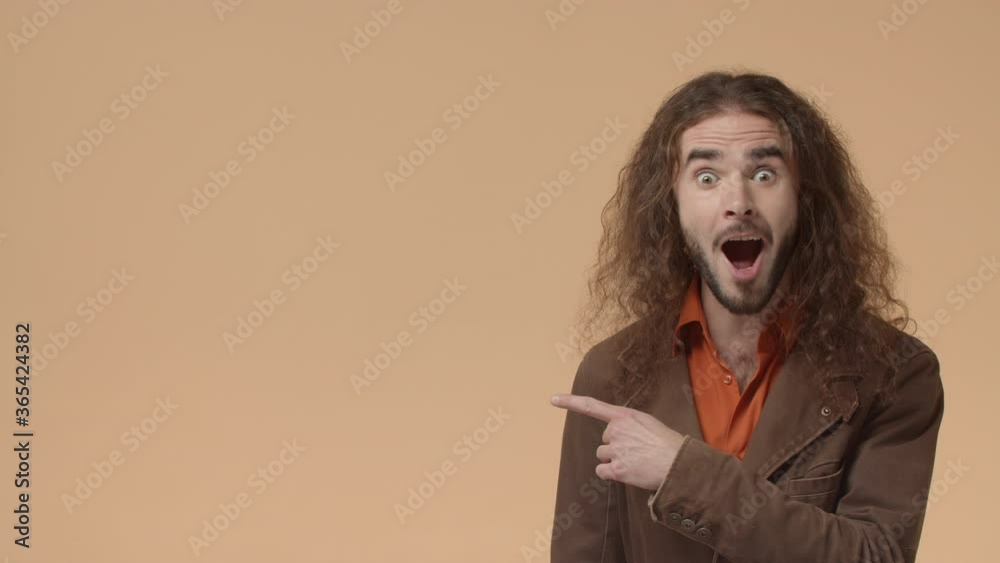 Big announcement, promo concept. Impressed and shocked, surpirsed bearded guy with long hair, look left with disbelief and then pointing at product advertisement excited and happy, beige background