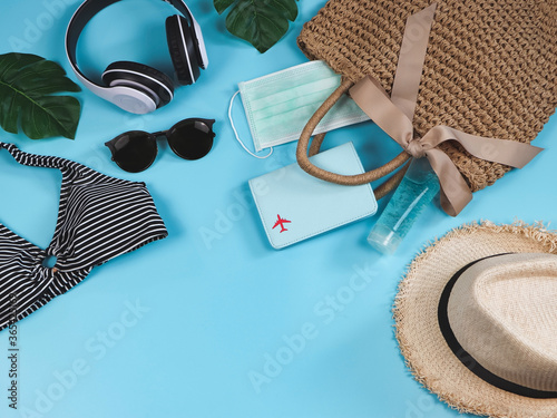 COVID-19 prevention , summer and new normal concept, top view of bikini swim wear and women's vacation accessories with passport surgical mask and alcohol gel in woven bag on blue background.