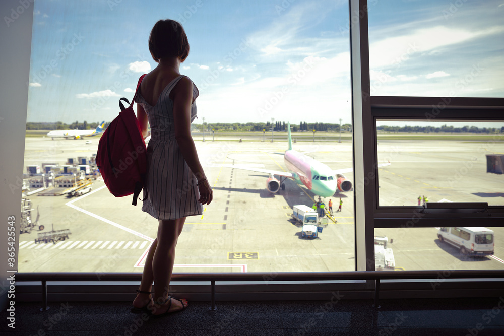 Obraz premium Caucasian woman standing inside airport and waiting her aircraft, looking through the window, airplane preparing for flight 