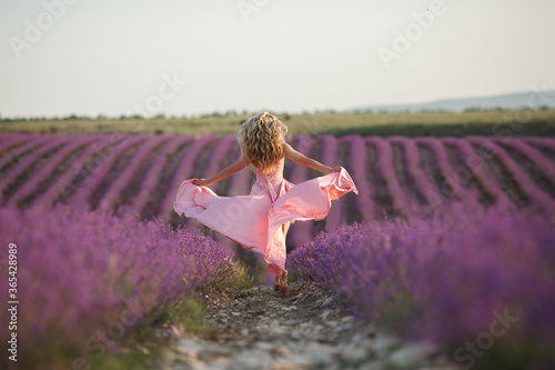 Fototapeta Naklejka Na Ścianę i Meble -  pretty blonde woman running away in lavender field. woman in long dress and straw hat having fun in flowers of lavender