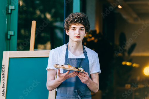 Young toughtful curly italian male waiter with food on tray serving at restaurant outdoor.Side jobs in bakery shop. Family business concept. photo