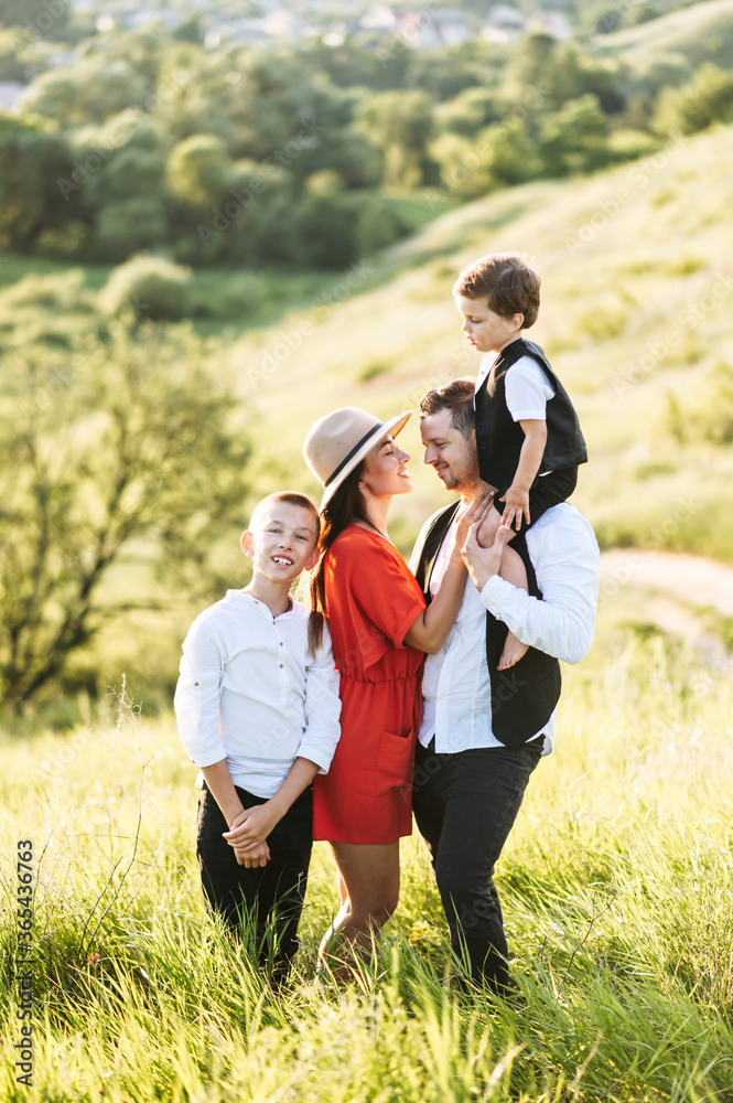 Longshot of a happy loving family standing on field in casual smart wear. Parents are caring looking on each other while the older son stands beside his family, the younger sits on father's shoulders Longshot of a happy loving family standing on field in casual smart wear. Parents are caring looking on each other while the older son stands beside his family, the younger sits on father's shoulders