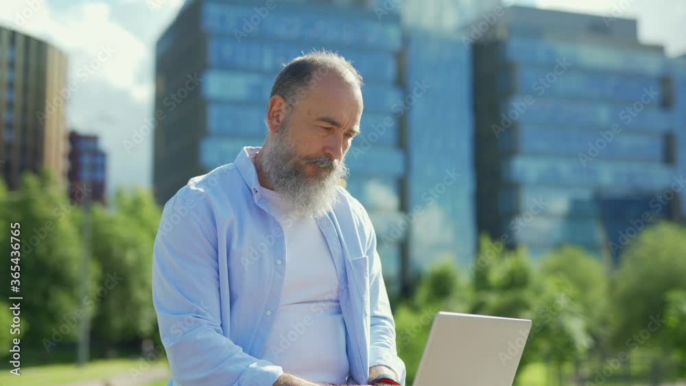 Zoom in medium shot of senior man with grey beard opening laptop, working on pc, thinking and closing computer sitting on bench outside on summer day