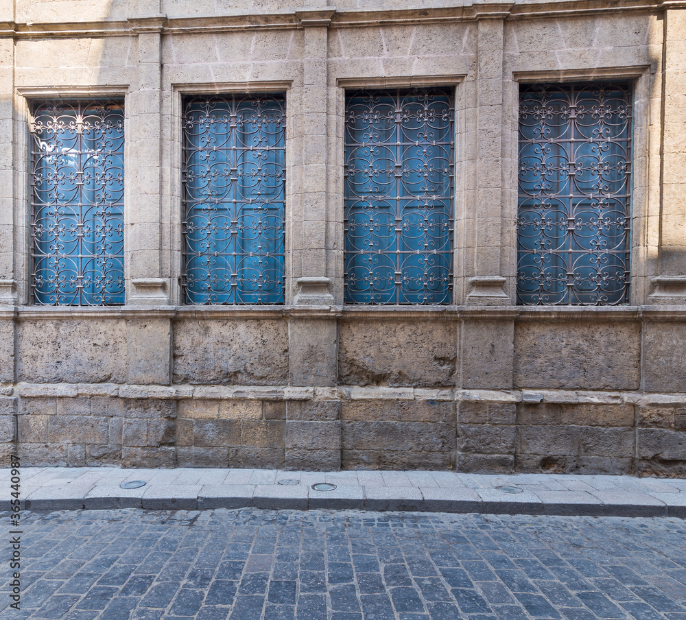 Row of four windows with ferforge wrought iron grill and closed blue wooden shutters on old stone bricks wall, and basalt tiles road Row of four windows with ferforge wrought iron grill and closed blue wooden shutters on old stone bricks wall, and basalt tiles road