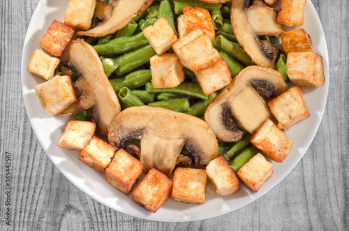 A delicious dish of green beans, fried mushrooms and diced cheese. Top view, part of a plate on a wooden background