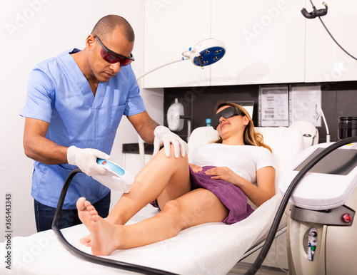 Professional Hispanic male cosmetologist performing laser hair removal for young female patient in clinic of esthetic cosmetology