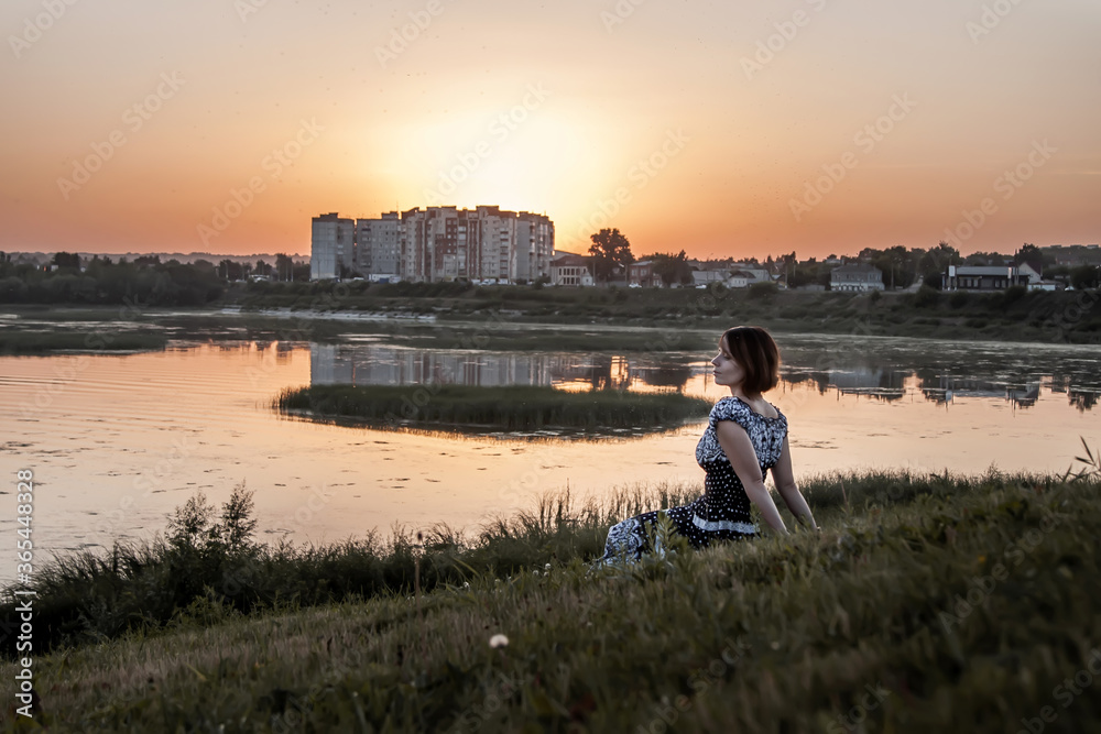 A girl in a dress sits on the river bank at sunset