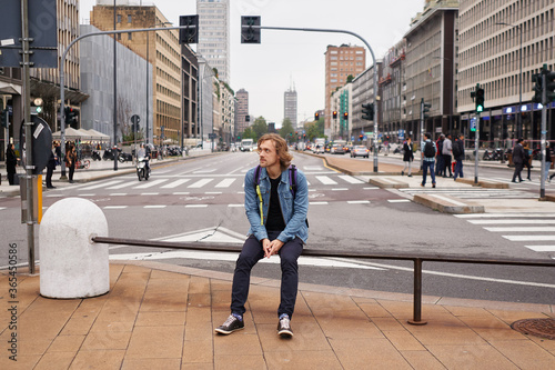 Young attractive man in casual wear and jeans jacket sitting on a fence with skyscrapers and big city background looking to the left side. Big city and traveling concept.