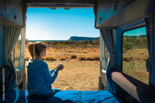Holidays in Australia. Woman having a coffee in the morning at the back side of a caravan, she wears casual clothes. Motorhome without logos. Vintage look. Kings Canyon, Northern Territory, Australia