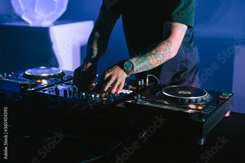Tattooed arms of a DJ mixing music at a party with his mixing board in a dark room with purple light.