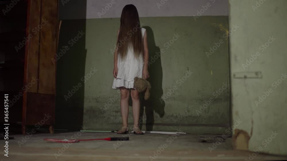 A demonic psycho child in a cursed abandoned house. A little girl dressed in a white dress with a bear toy looks scared at the 4K camera footage A demonic psycho child in a cursed abandoned house. A little girl dressed in a white dress with a bear toy looks scared at the 4K camera footage