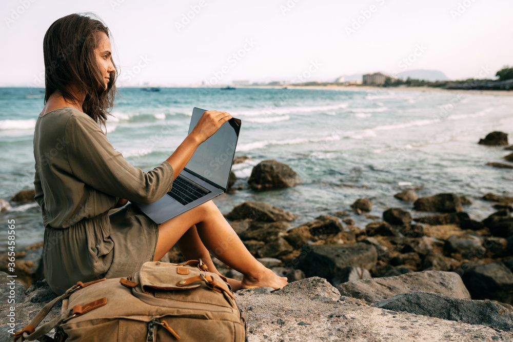 Young girl freelancer works sitting on a rocky beach by the sea at sunset, working in a non-office. The concept of remote work