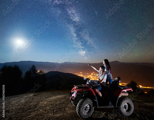 Young couple man and woman tourists riding atv quad motorbike on the top of mountain. Man pointing at beautiful night sky full of stars  full moon  Milky way  luminous village on background