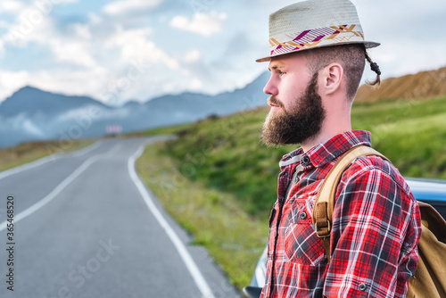 Handsome young man in a straw hat and a plaid shirt, stands on the road, against the backdrop of beautiful mountains. The concept of freedom and hitchhiking © Валентина Баранова