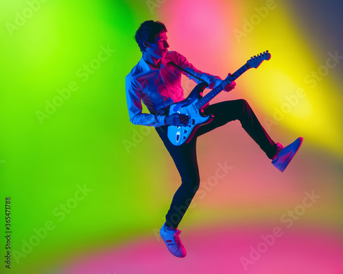Jump high. Young caucasian inspired and expressive musician, guitarist performing on multicolored background in neon. Concept of music, hobby, festival, art. Joyful artist, colorful, bright portrait.