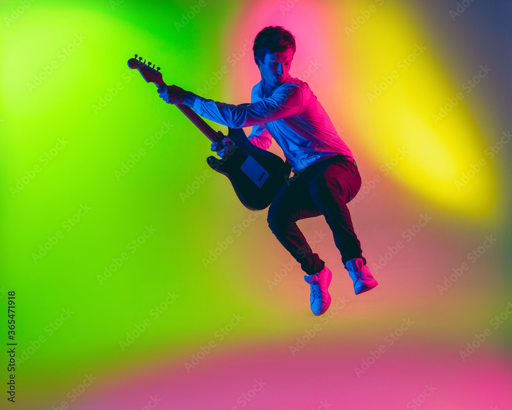 Jump high. Young caucasian inspired and expressive musician, guitarist performing on multicolored background in neon. Concept of music, hobby, festival, art. Joyful artist, colorful, bright portrait.
