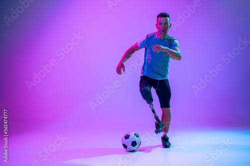 Fototapeta Naklejka Na Ścianę i Meble -  Athlete with disabilities or amputee on gradient studio background in neon. Professional male football player with leg prosthesis training in studio. Disabled sport and healthy lifestyle concept.