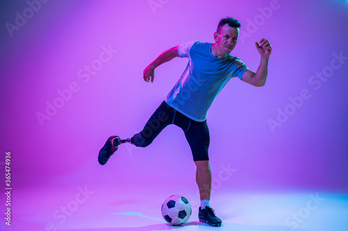 Athlete with disabilities or amputee on gradient studio background in neon. Professional male football player with leg prosthesis training in studio. Disabled sport and healthy lifestyle concept.