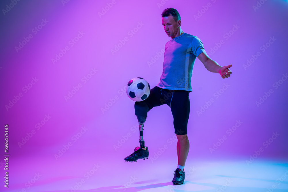 Athlete with disabilities or amputee on gradient studio background in neon. Professional male football player with leg prosthesis training in studio. Disabled sport and healthy lifestyle concept.