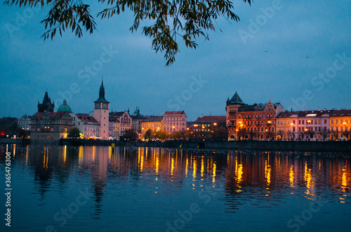 An early evening in Prague during autumn with incandescent lights illuminating the city and reflections over the Vltava river with several ducks floating in it.