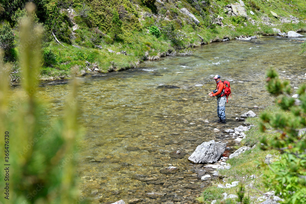 fly fisherman trout fishing with a hiking backpack and an orange jacket in the high mountains in summer