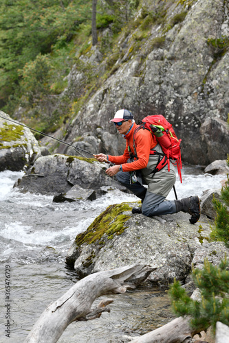 fly fisherman trout fishing with a hiking backpack and an orange jacket in the high mountains in summer