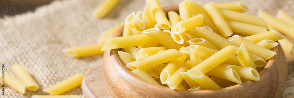 Penne pasta. Raw Penne Pasta in a wooden bowl on a brown wooden table. Banner with space for text Penne pasta. Raw Penne Pasta in a wooden bowl on a brown wooden table. Banner with space for text