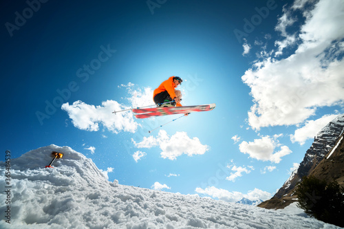 A young stylish man in sunglasses and a cap performs a trick in jumping with a kicker of snow against the blue sky and the sun on a sunny day. The concept of park skiing in winter or spring