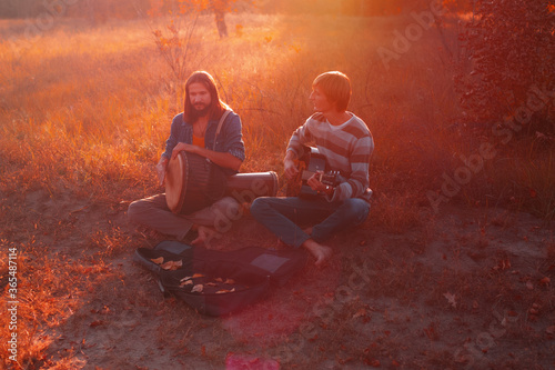 Two hippie men playing djembe drum and acoustic guitar in the autumn in the forest at sunset.