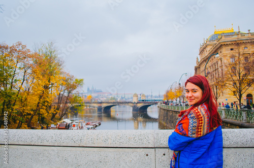 A redhead brazilian woman in her 20s in front of a bridge in Prague, Czechia and smiling to the camera.