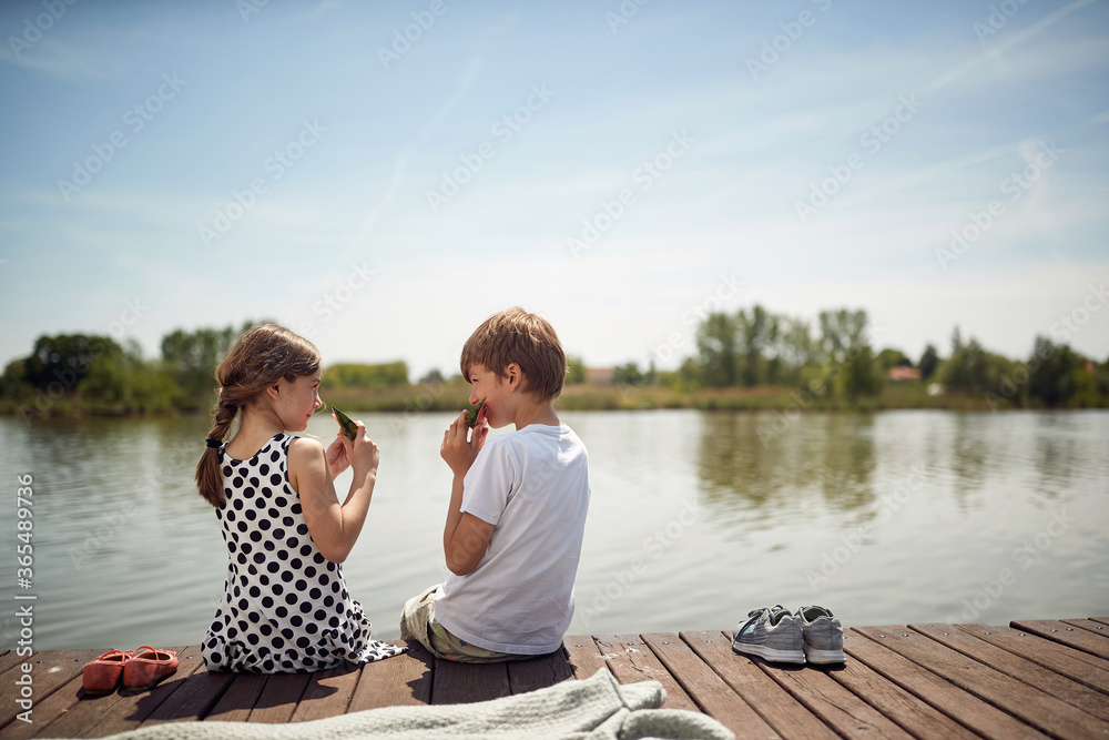 Children  on wooden near pond eating fresh watermelon.