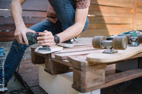 Close-up view of sanding and polishing wooden board. Cropped man using sanding machine on wooden board on table with skateboard.