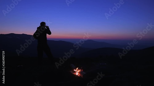 A man drinks a hot drink at night by a campfire in a tourist camp on a mountain pass. A tourist admires the beautiful views of the mountains after sunset. Hiking in the mountains.