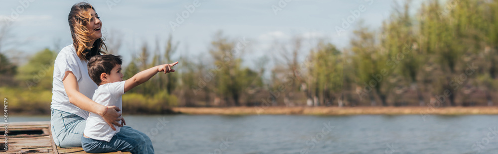 Panoramic concept of cute kid pointing with finger while sitting near happy mother and lake - obrazy, fototapety, plakaty 