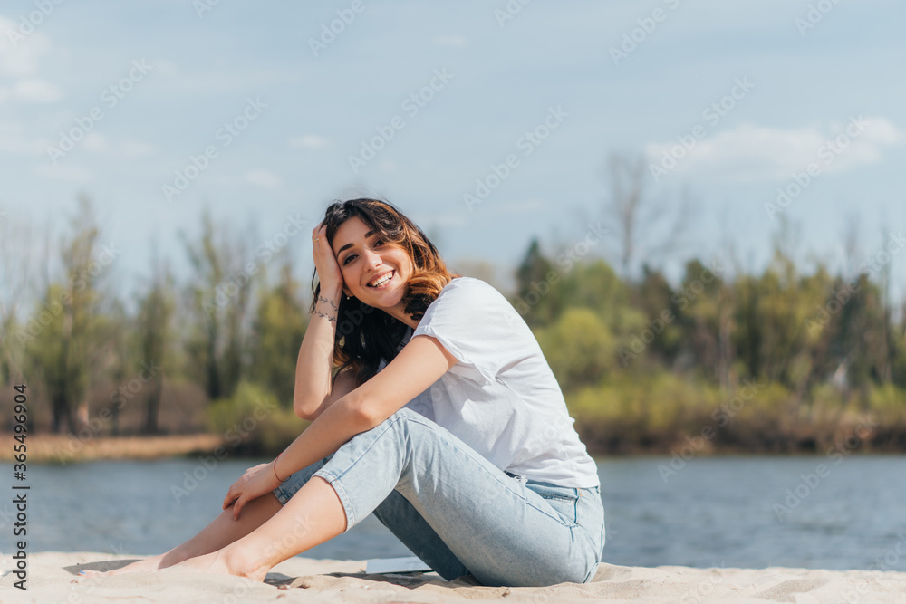 cheerful woman sitting on sand near river and looking at camera
