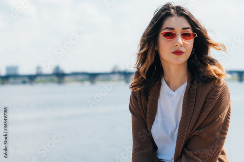 trendy woman in stylish sunglasses looking at camera near lake