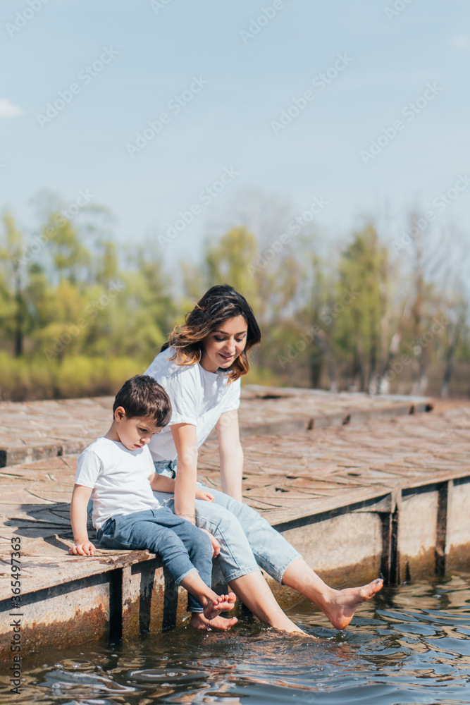 Barefoot mother and cute son sitting near lake