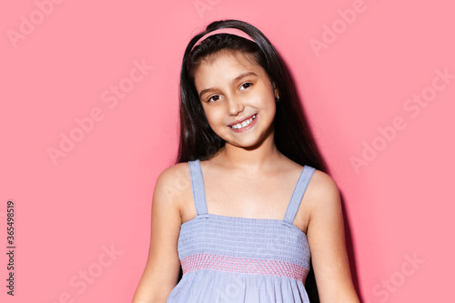 Studio portrait of happy little brunette child girl on background of pastel pink color. Wearing blue dress and headband.