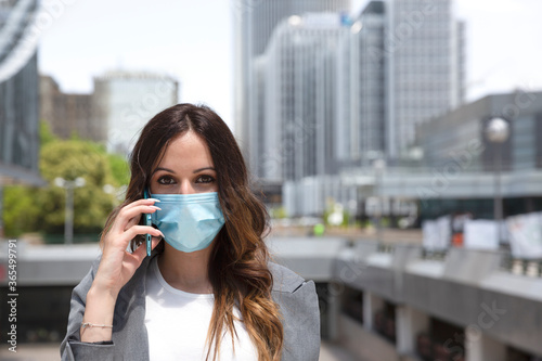 Close-up of young entrepreneurial woman talking through her smartphone outside a business area. She is wearing a face mask. Selective focus. Space for text. Business and new normal concept. 