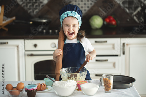 Cute little girl in apron and chef hat is flattening the dough using a rolling pin, looking at camera and smiling while baking