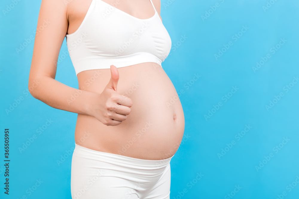 Cropped shot of positive pregnant woman in white underwear showing thumb up cool gesture over her baby bump at blue background. Happy maternity. Copy space Cropped shot of positive pregnant woman in white underwear showing thumb up cool gesture over her baby bump at blue background. Happy maternity. Copy space