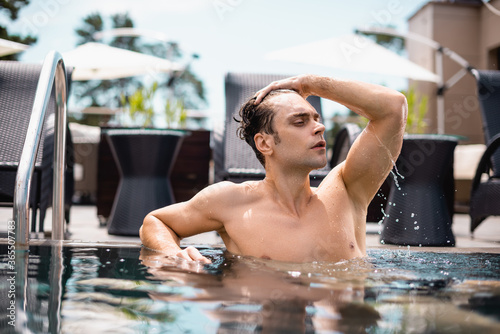 shirtless man looking away and touching wet hair in swimming pool